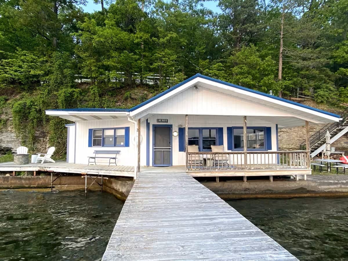 The white cottage with blue trim seen from the end of its private dock, with clear green water on both sides of the dock walkway and a wooded bluff rising behind.