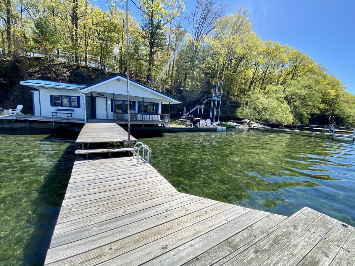 Wide view along the weathered dock toward the cottage, with shallow clear water showing the shale bottom and bare spring trees on the hillside.