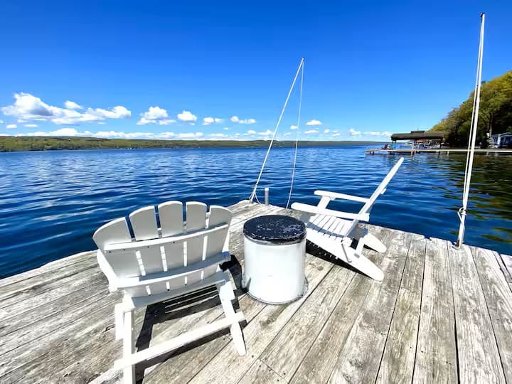 Two white Adirondack chairs on the dock on a bright day, deep blue water and the far shore in the distance.