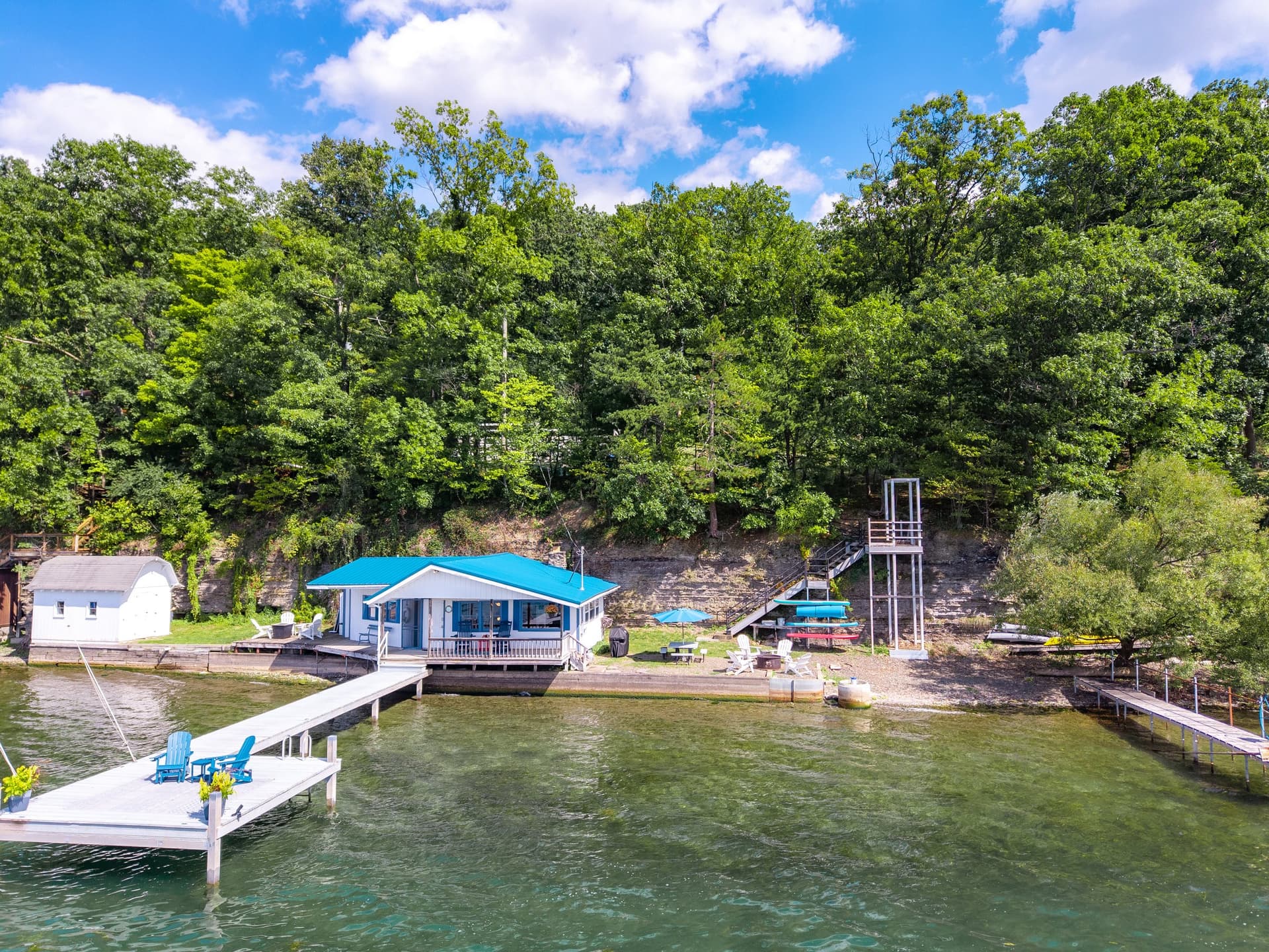 The dock and shoreline from above the water on a bright day, chairs at the dock head, the cottage under its blue roof, and the tree-covered slope running along the shore.