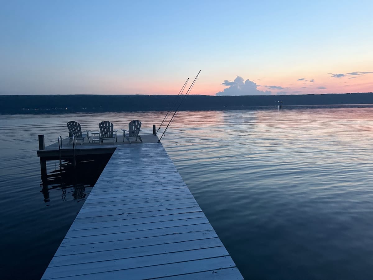 The dock at dusk leading out to three chairs at its end, with glassy water reflecting a pink and pale blue sky.