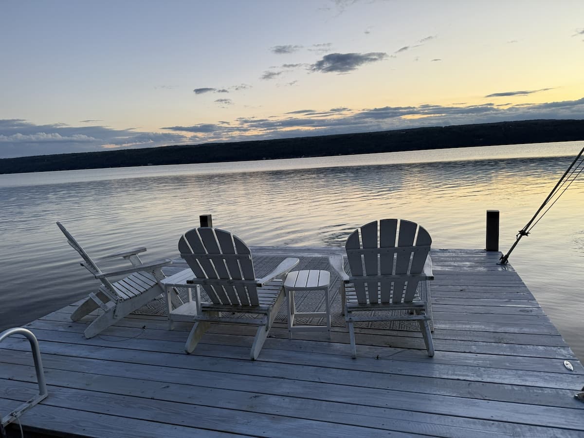 Three Adirondack chairs and a small side table on the dock at sunset, facing still water and a gold and blue sky over the far shore.