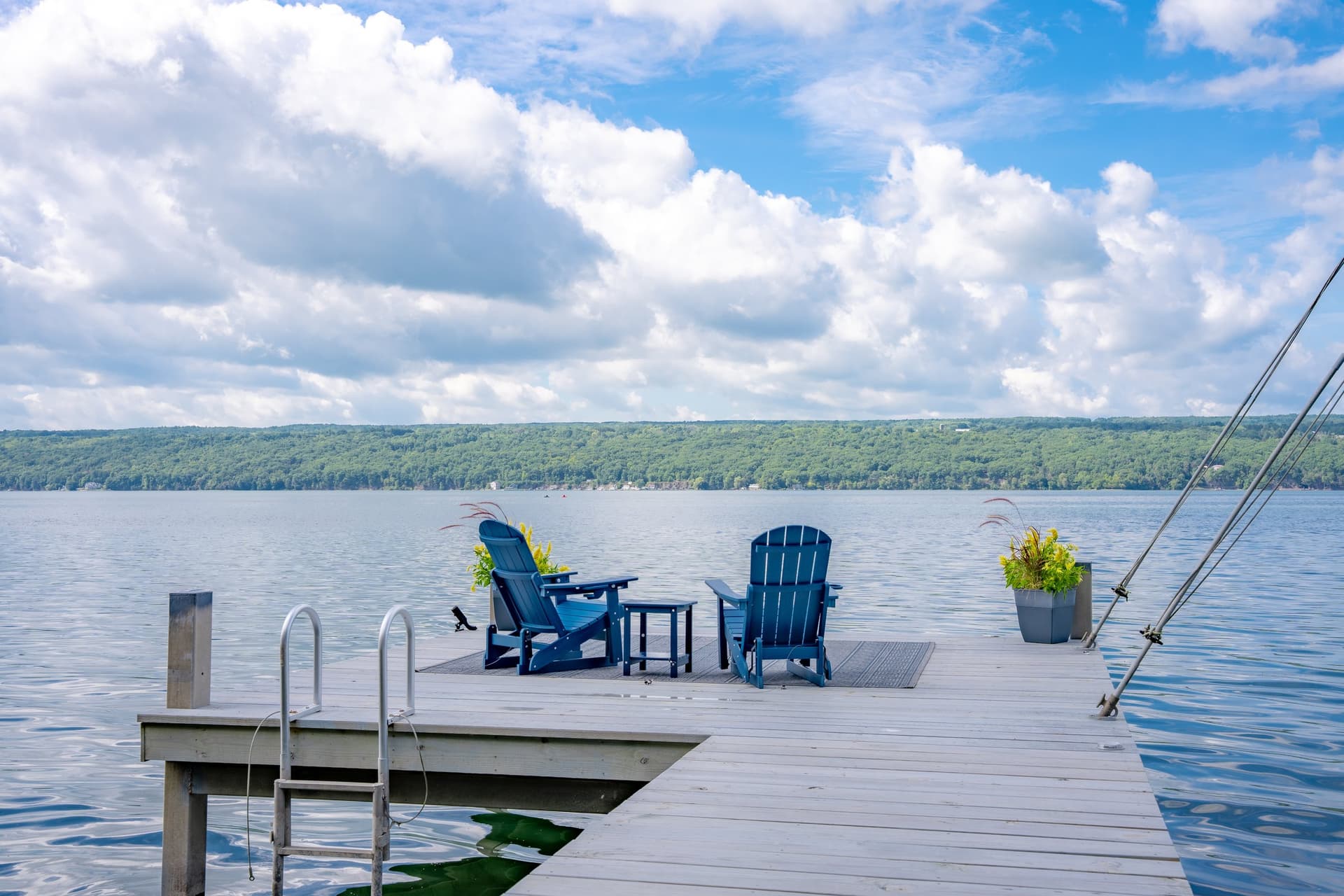 Two blue Adirondack chairs and a small table at the end of the dock, with a swim ladder at the edge, a potted plant, and open water running to the far shore under a tall cloudy sky.