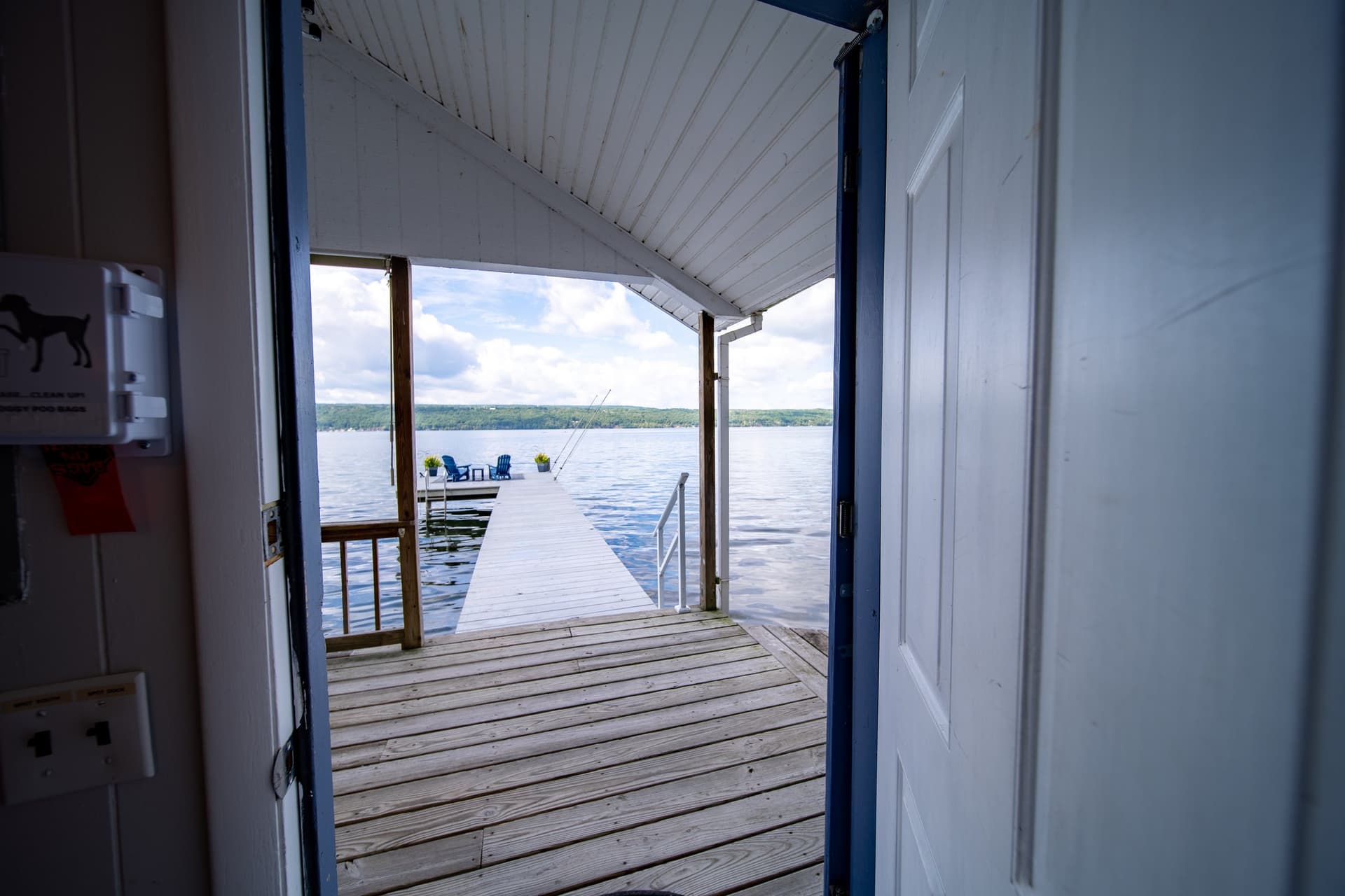 The open lakeside door of the cottage framing the dock beyond it, boards running straight out to chairs at the far end and water on both sides.