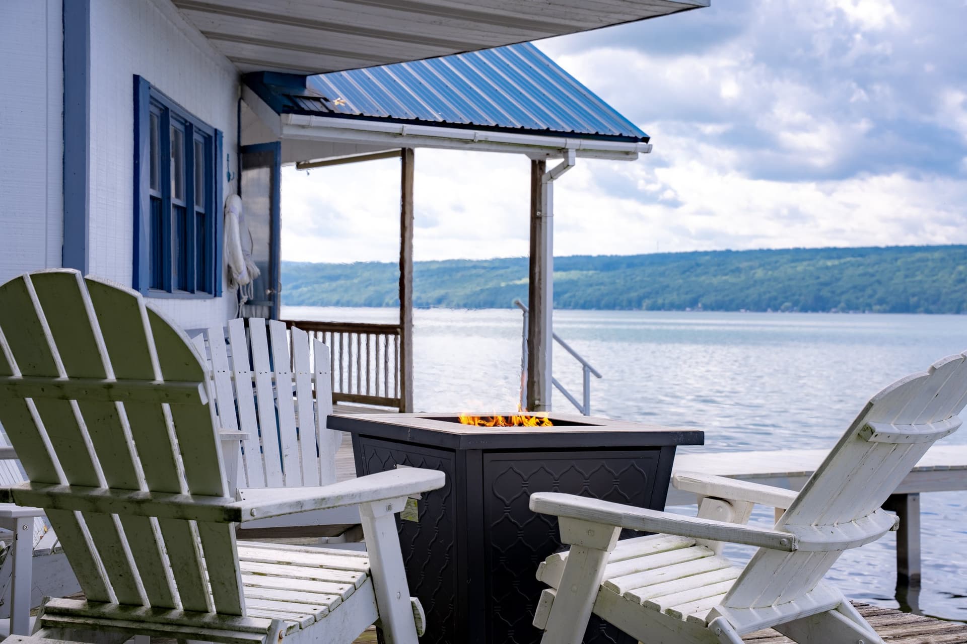 White Adirondack chairs pulled around a lit fire table beside the cottage porch, with the blue roof overhead and the lake and far shore beyond.
