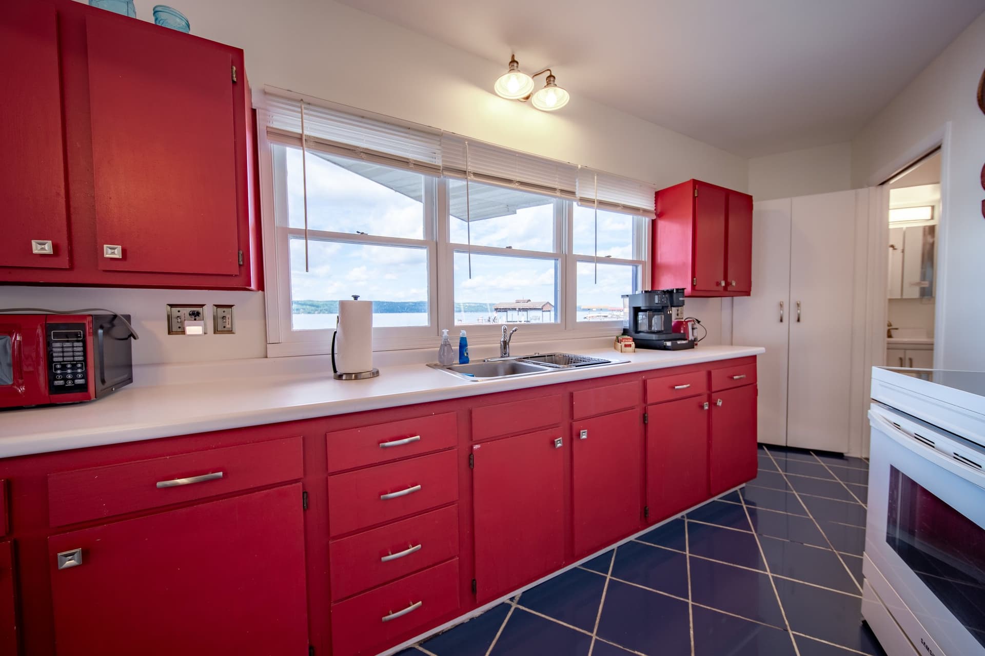 The galley kitchen along one wall with red cabinets, white counters, an electric range, and a run of windows above the counter showing the lake.