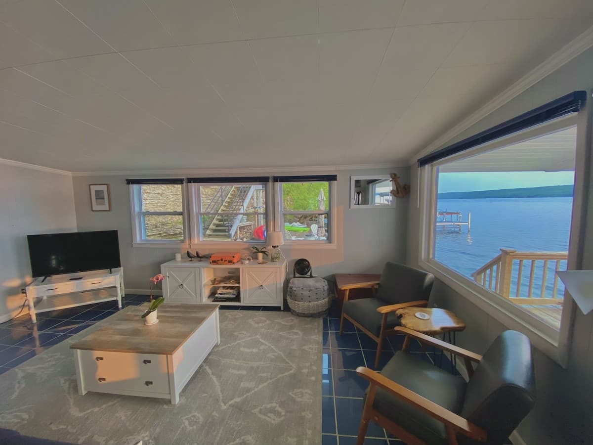 The living room with a large picture window looking directly onto the lake and the deck rail, cobalt tile floor, grey armchairs and a low white coffee table.