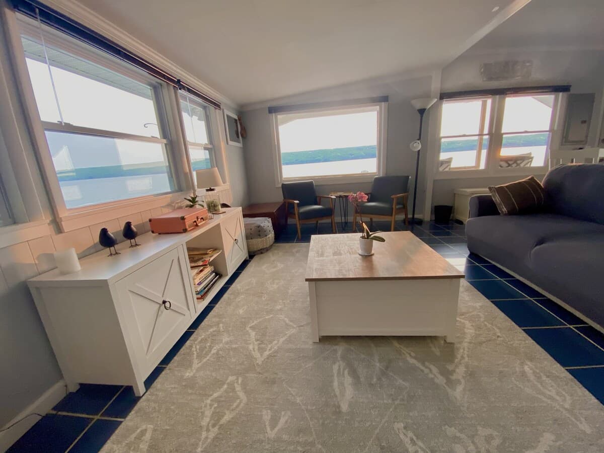 Living room looking along a wall of windows with the lake visible through each one, a navy sofa, white storage cabinets and a patterned grey rug over cobalt tile.