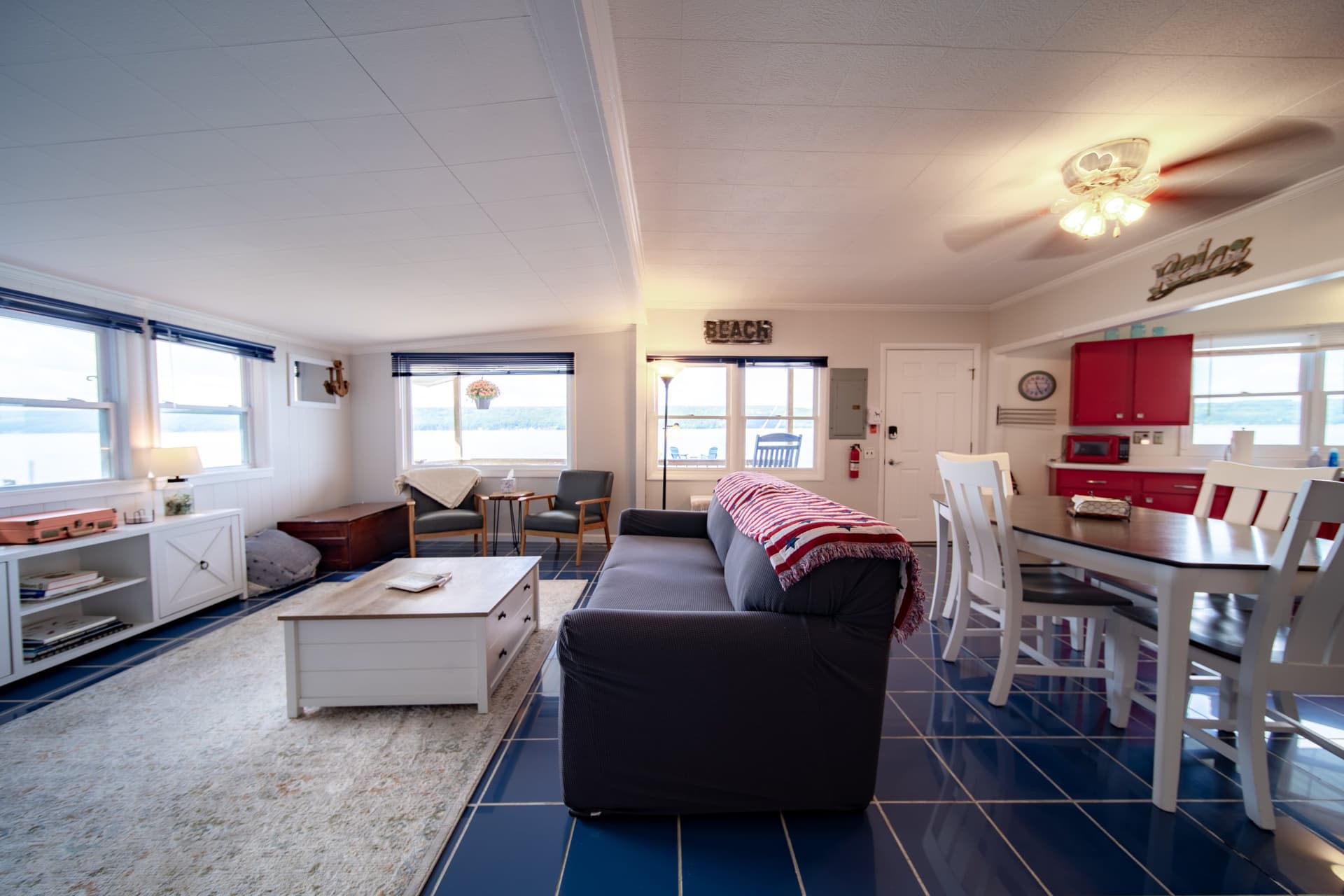 The open living and dining space end to end: sofa and coffee table, the white dining table and chairs, the red kitchen cabinets beyond, and lake through the windows along the whole wall.