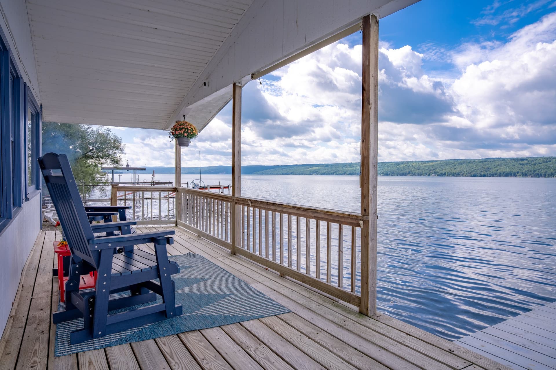 A navy rocking chair and a small red table on the covered porch, the plank deck running to a rail, and the lake and far shore filling the right of the frame.