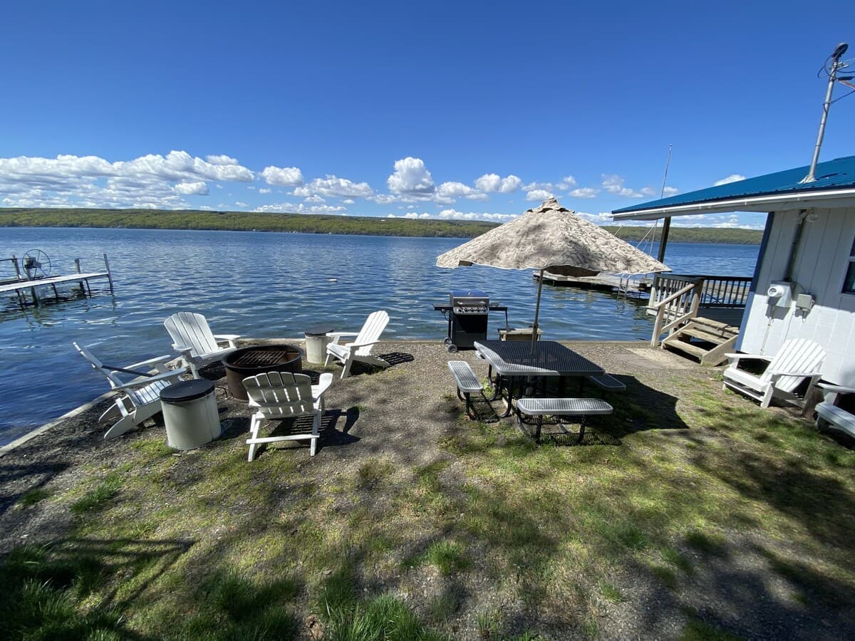 Shore terrace beside the cottage with Adirondack chairs around a fire ring, a picnic table under a canvas umbrella, and a gas grill, looking north up the open lake.